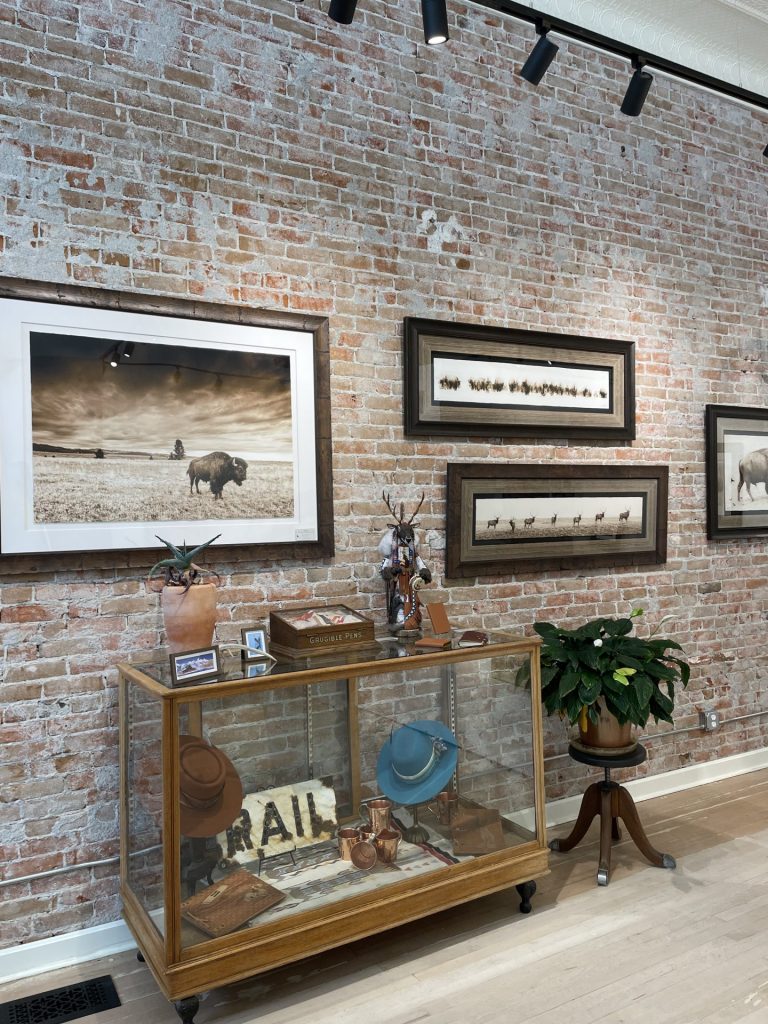 Several pieces of artwork displayed on a brick wall plus more artwork, artifacts and merchandise inside and on top of a display case at the Medicine Bird Gallery
