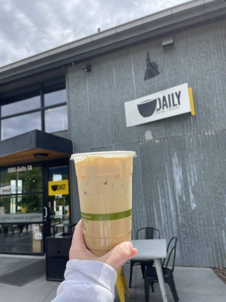 Someone holding a beverage in a to-go cup in front of 'the daily' coffee building and signage