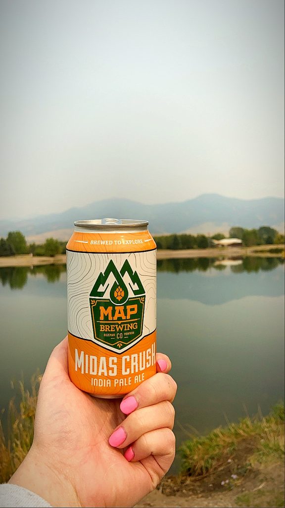 Someone holding a can of beer outside of map brewing with pond and mountains in the background on a hazy day